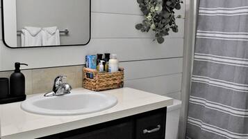 Een badkamer met een witte wastafel, een zwarte kraan en een spiegel erboven. Op het aanrecht staat een rieten mand met toiletartikelen, en er hangt een groene plant aan de muur.