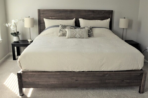 A large bed with a dark headboard, two bedside tables with lamps, and a potted plant on one side.