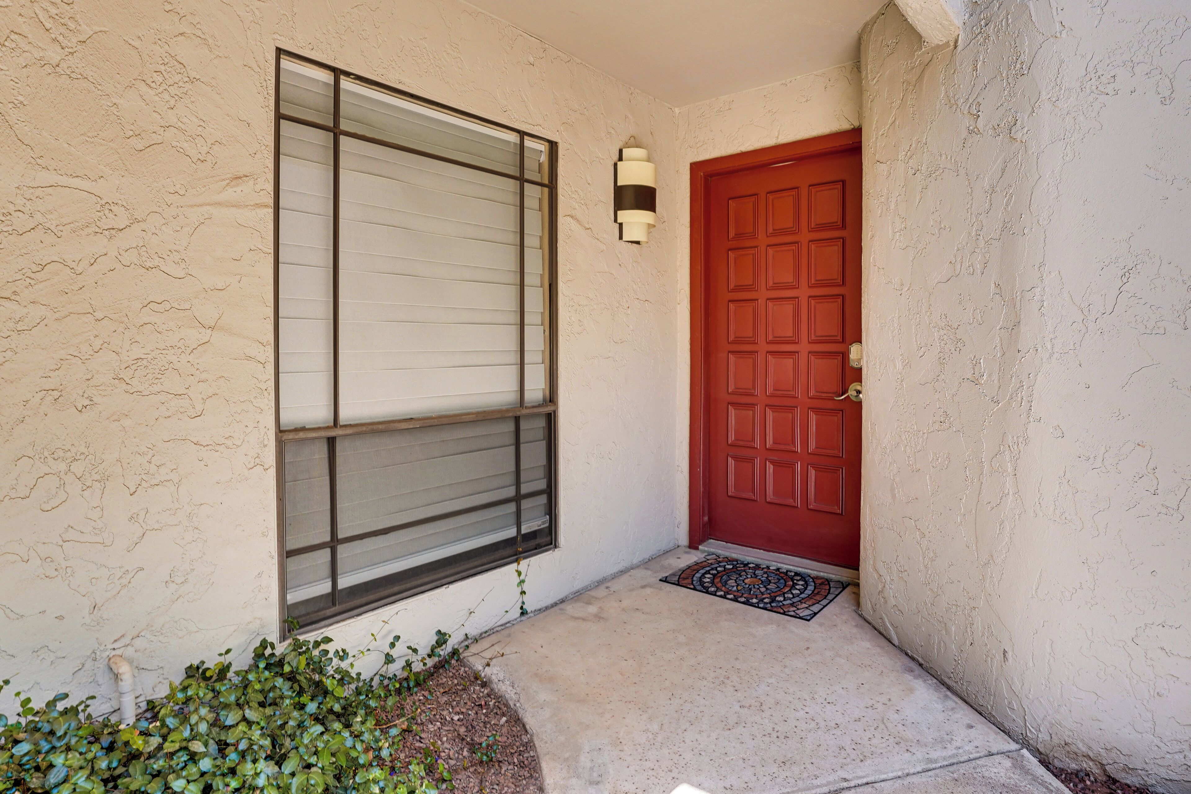 A building entrance with a red door, a window with blinds, and a small garden area.