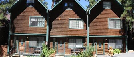 A row of log cabin-style buildings with green trim and wooden exteriors, featuring multiple windows and a front porch with a bench.