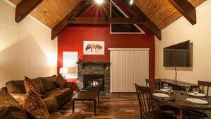 A cozy living room with a fireplace, wooden ceiling beams, and a dining area set for four.