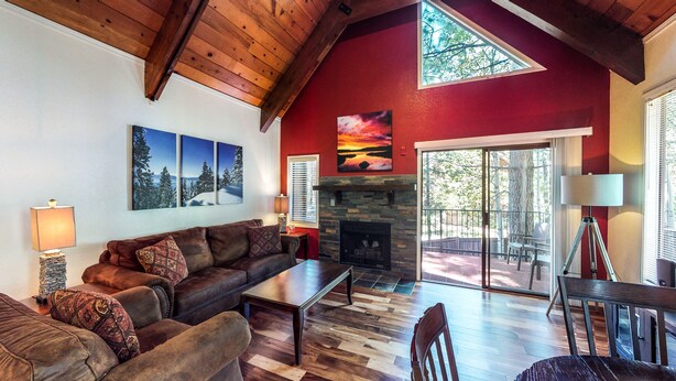 A cozy living room with a fireplace, wooden ceiling beams, and a large window overlooking a deck.