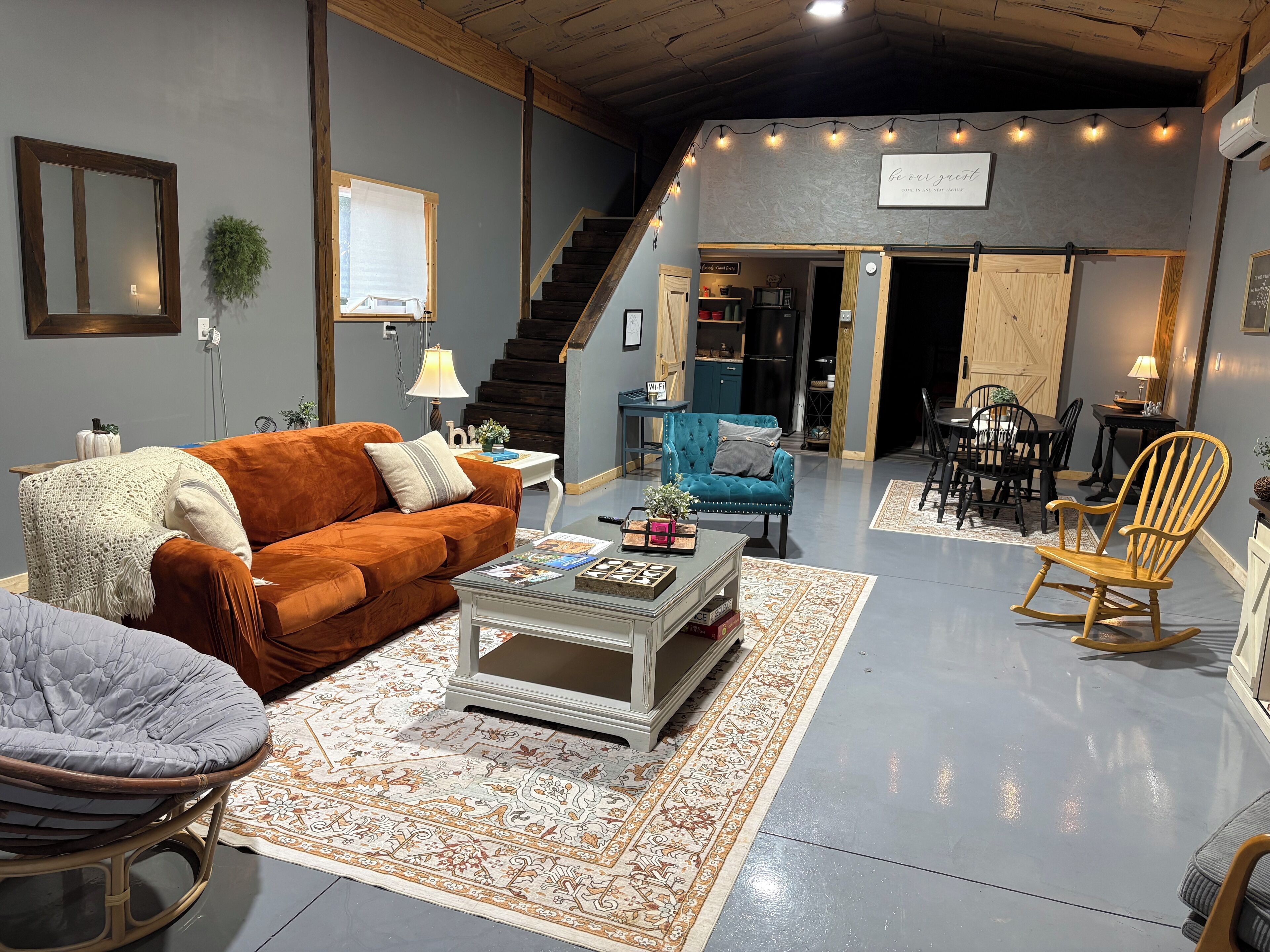Un salon spacieux avec un grand canapé, une table basse et un fauteuil à bascule. Un escalier mène à un autre niveau, et on aperçoit un coin cuisine au fond de la pièce.