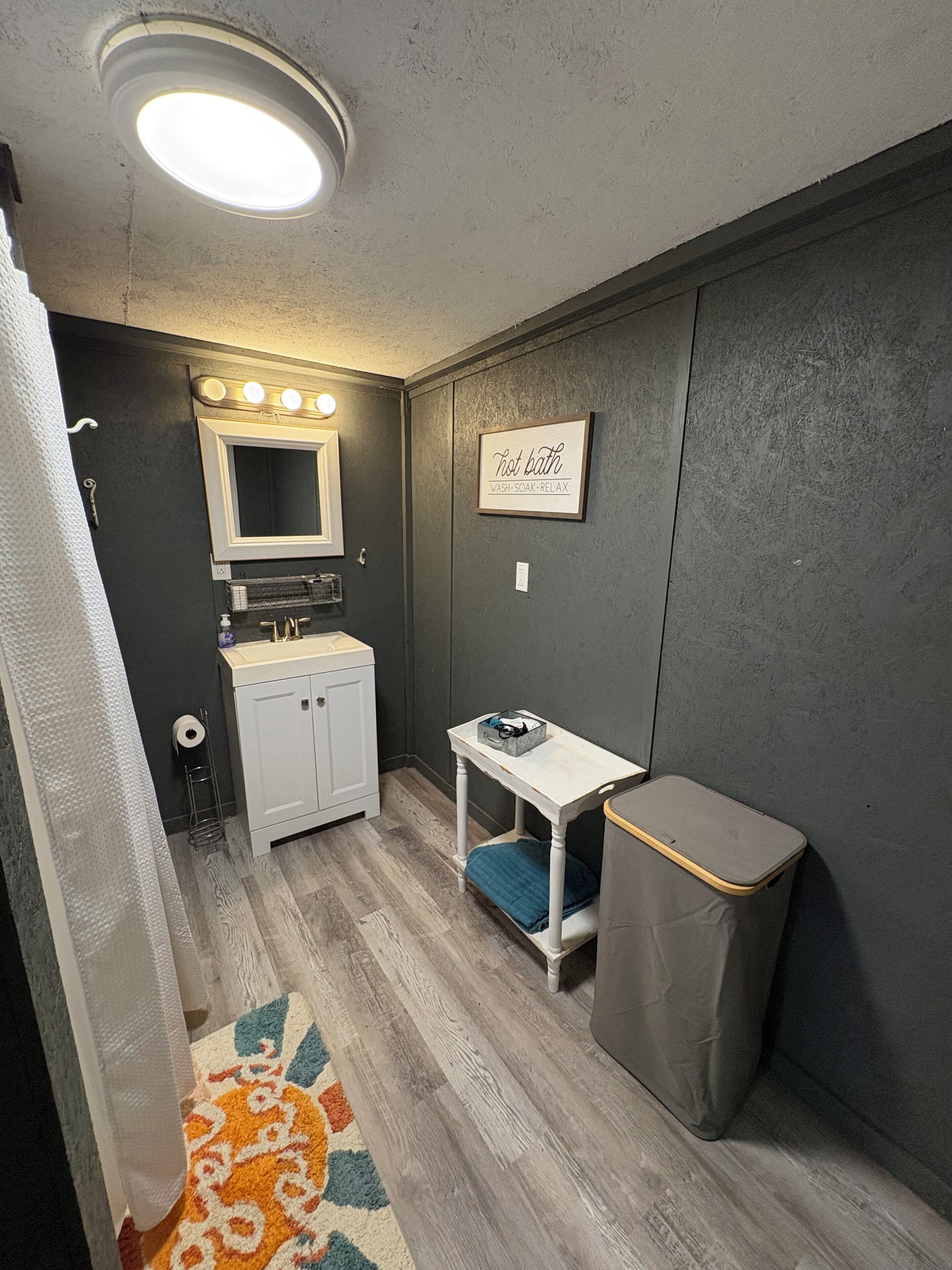 Een kleine badkamer met een wastafel, spiegel en een tafeltje met een afvalbak.