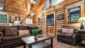 Ein Interieur einer Blockhütte mit Holzbalkendecke, zwei Ledersofas mit Kissen, einem Couchtisch und einem roten Teppich mit Muster.