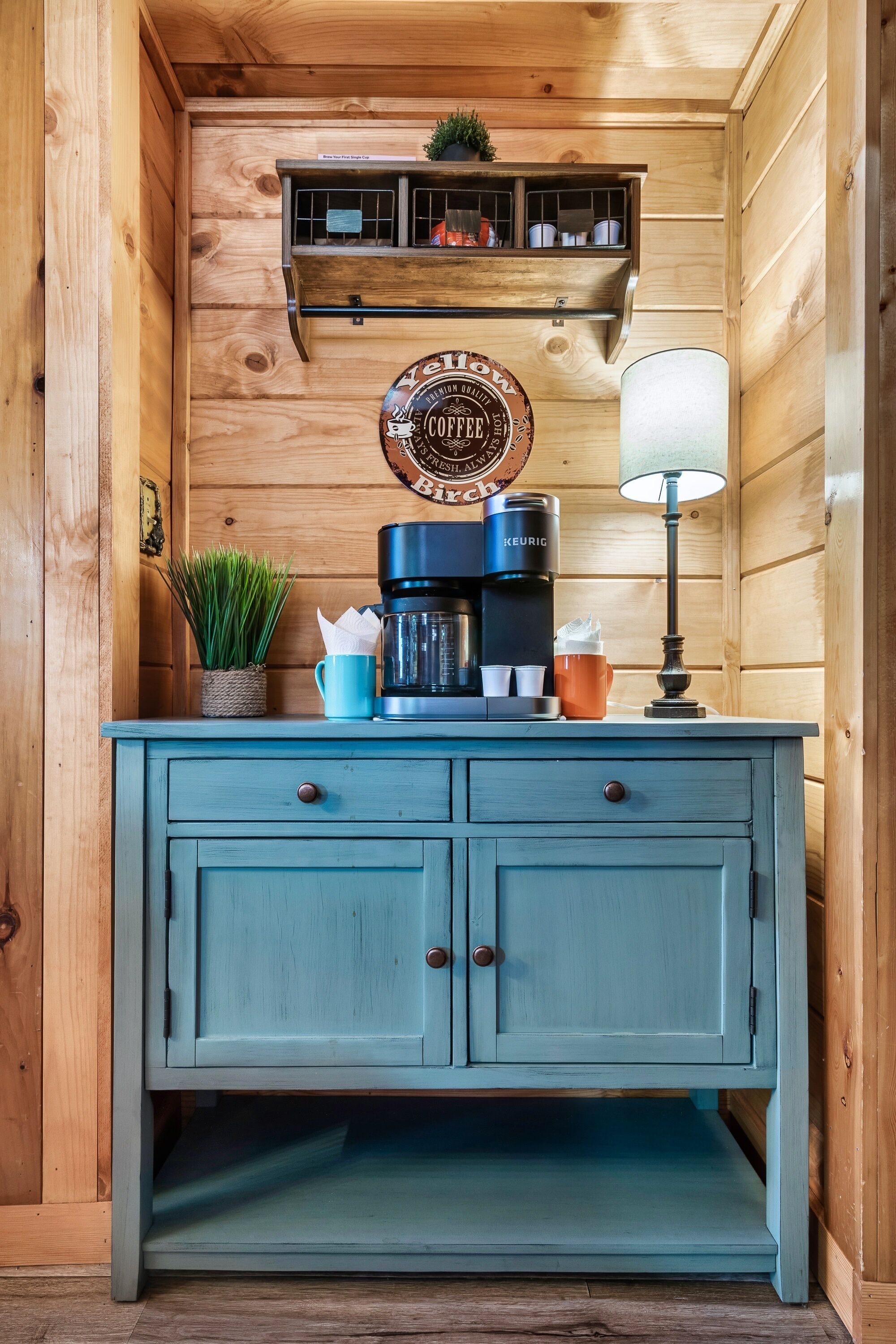 A wooden cabin interior with a blue cabinet, a coffee maker, a lamp, and a shelf with kitchen items.
