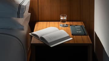 A neatly made bed with white linens and a black headboard, a bedside table with a book, a glass of water, and a black notebook.