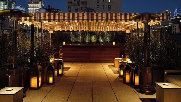 An outdoor seating area with a wooden pergola, ambient lighting, and a view of the city skyline at night. - The Moore, a Member of Design Hotels (New York)