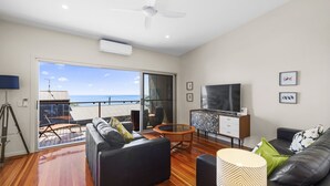 A modern living room with a large sliding glass door leading to a balcony, a flat-screen TV, and a wooden coffee table. - Rock Ledge - Woolgoogla Headland (Woolgoolga)