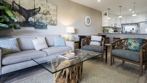 A modern living room with a glass coffee table, a sofa, and a dining area with wooden chairs. - *25% Off-Steps to Beach-AC in Every Room-Remodeled (Kihei)
