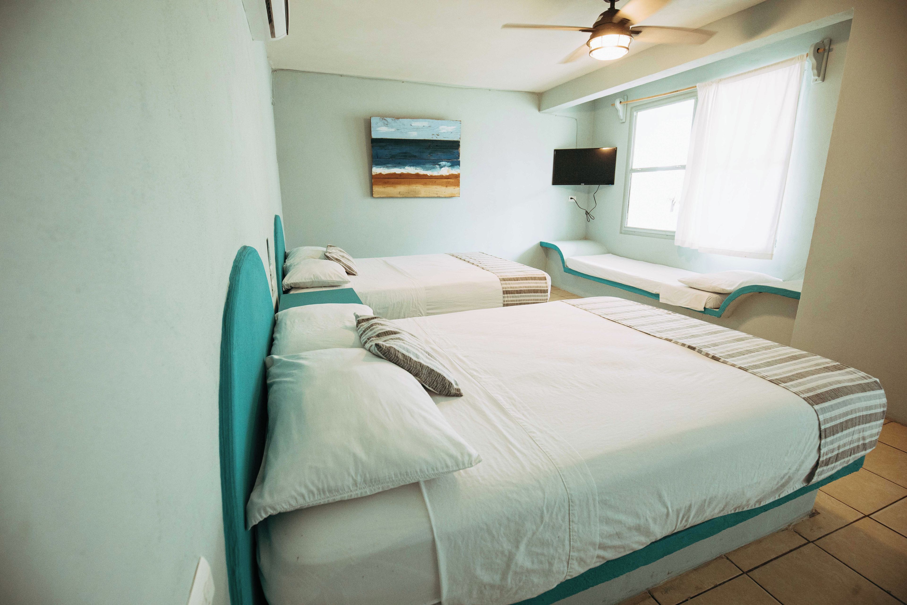 A room with two beds, a ceiling fan, a painting, a TV, and a window with curtains.