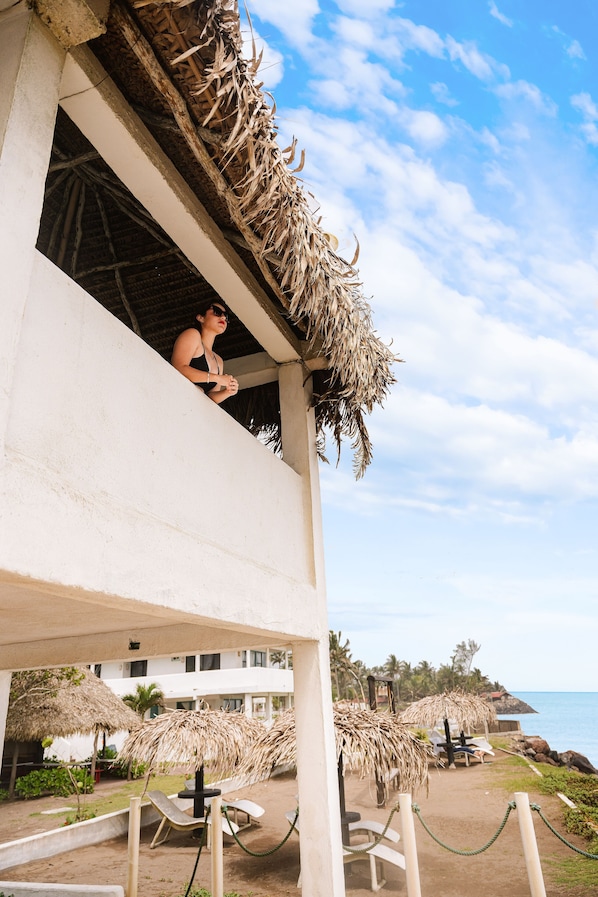 A beachside hotel with a balcony overlooking the ocean, thatched umbrellas, and lounge chairs.