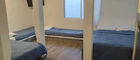 Una habitación con tres camas dispuestas en forma de U, una ventana que deja entrar luz natural, y piso de madera.