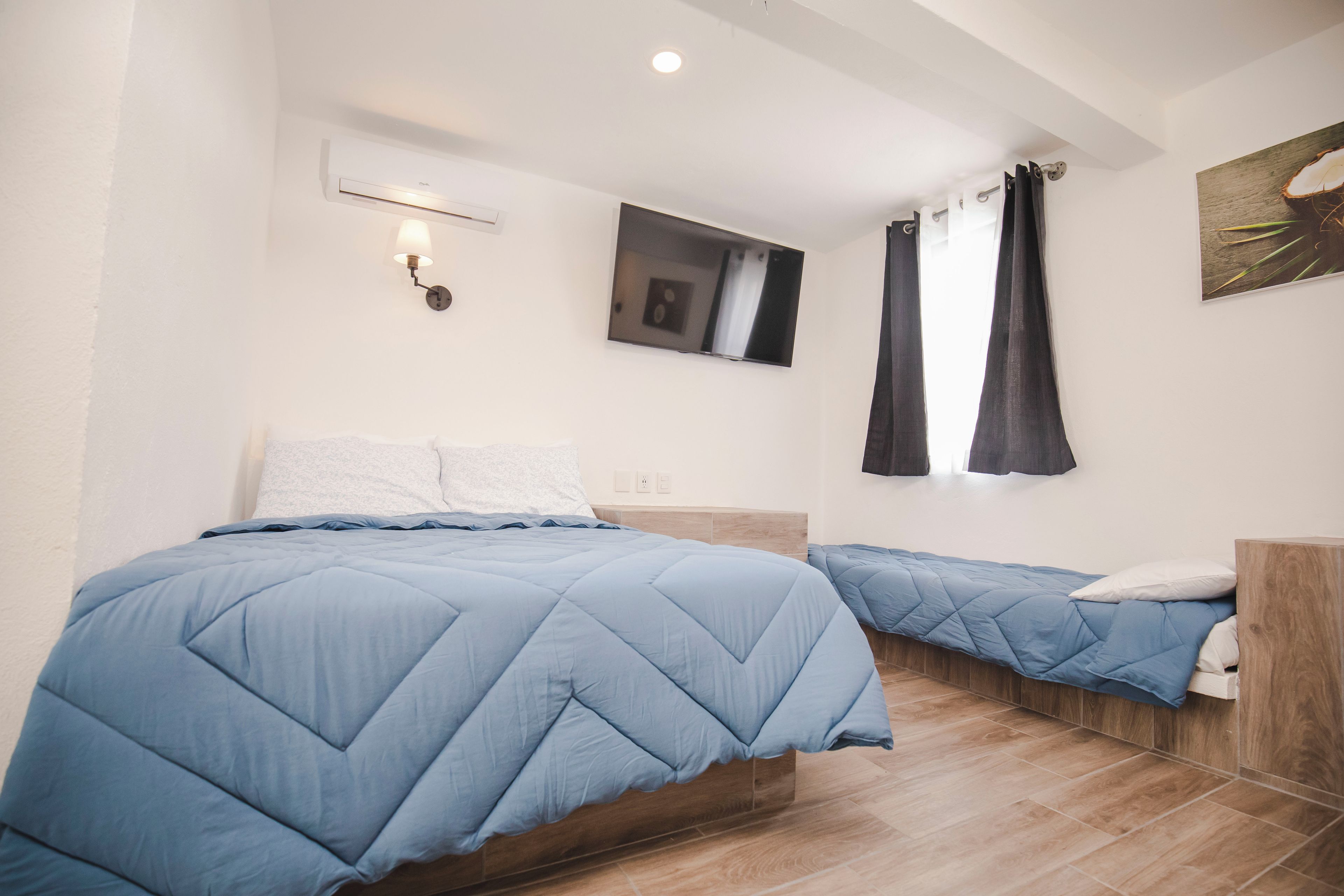 Un dormitorio con cama, mesita de noche, televisor en la pared y ventana con cortinas.