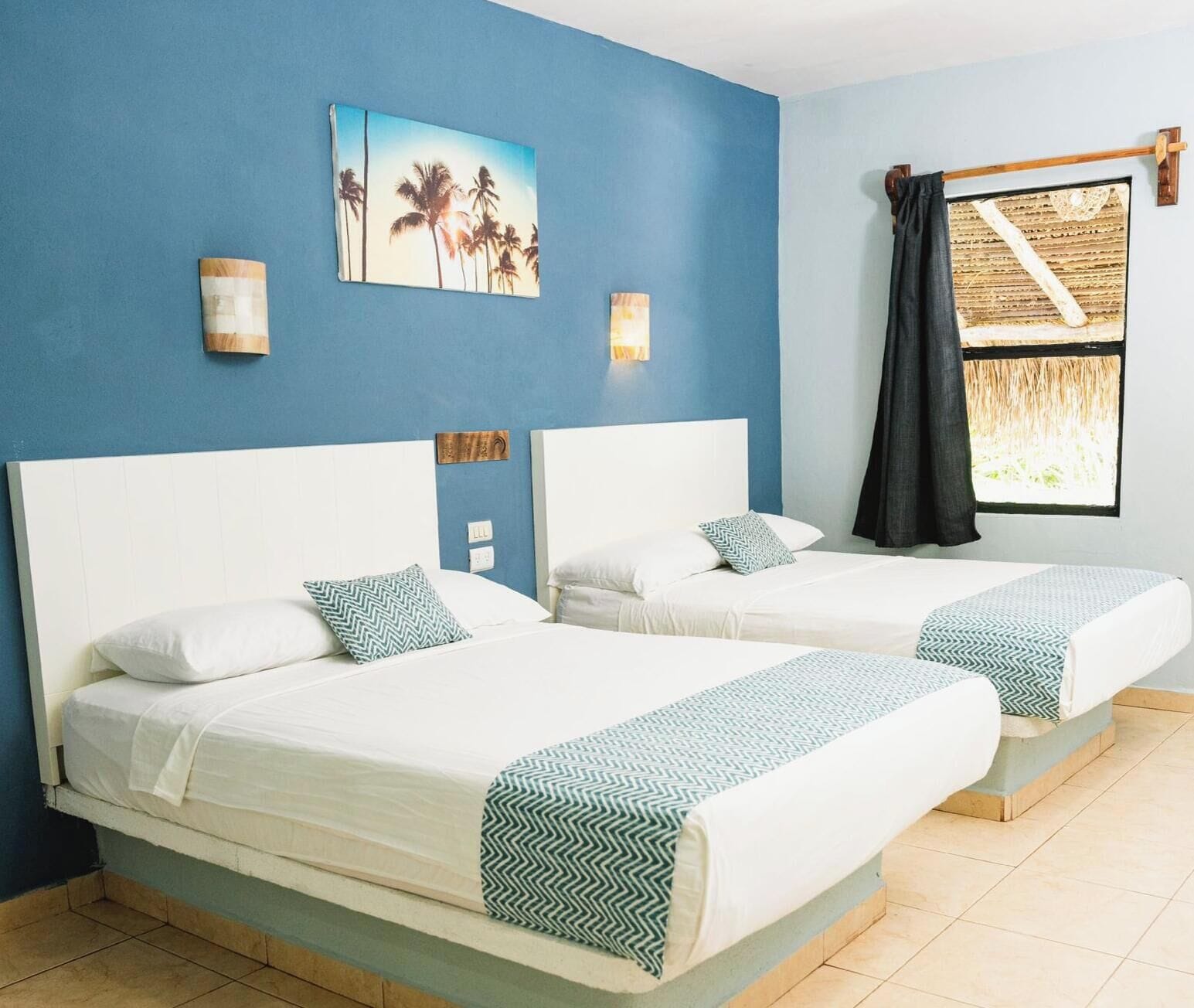 A hotel room with two beds, a blue wall, and a window with a curtain.