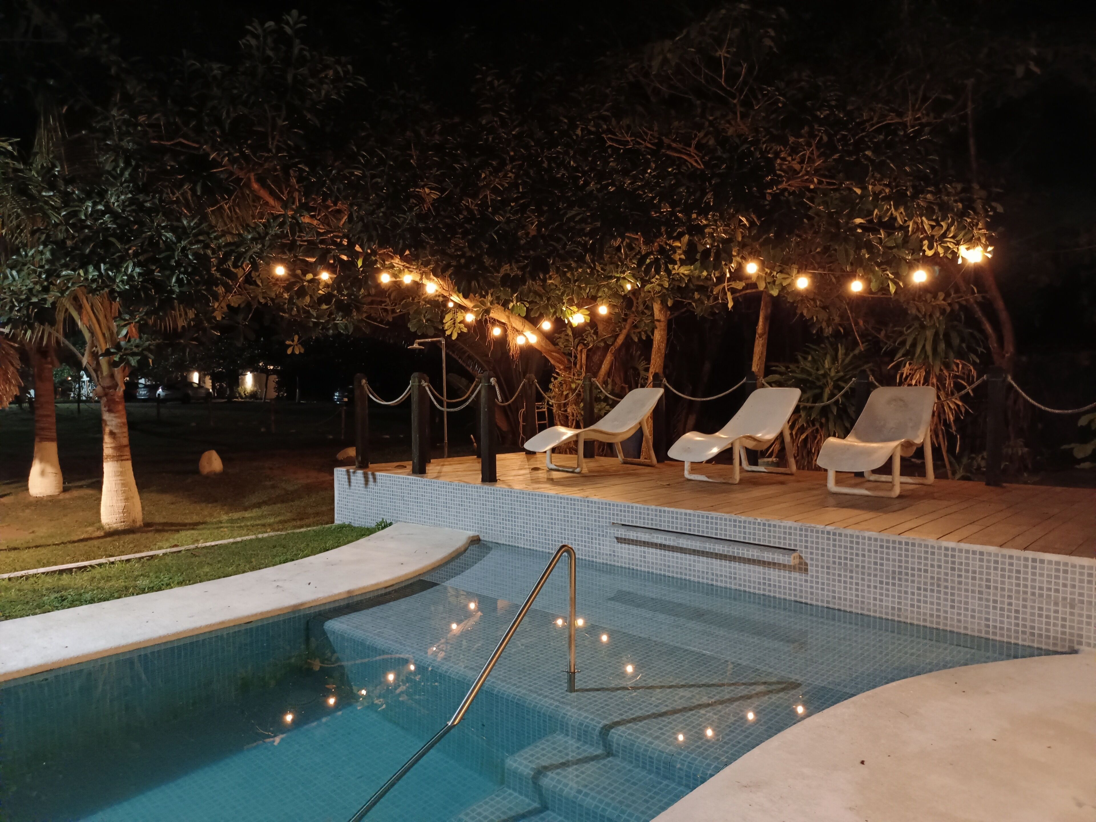 Un área de piscina con sillones de descanso e iluminación con luces colgantes.