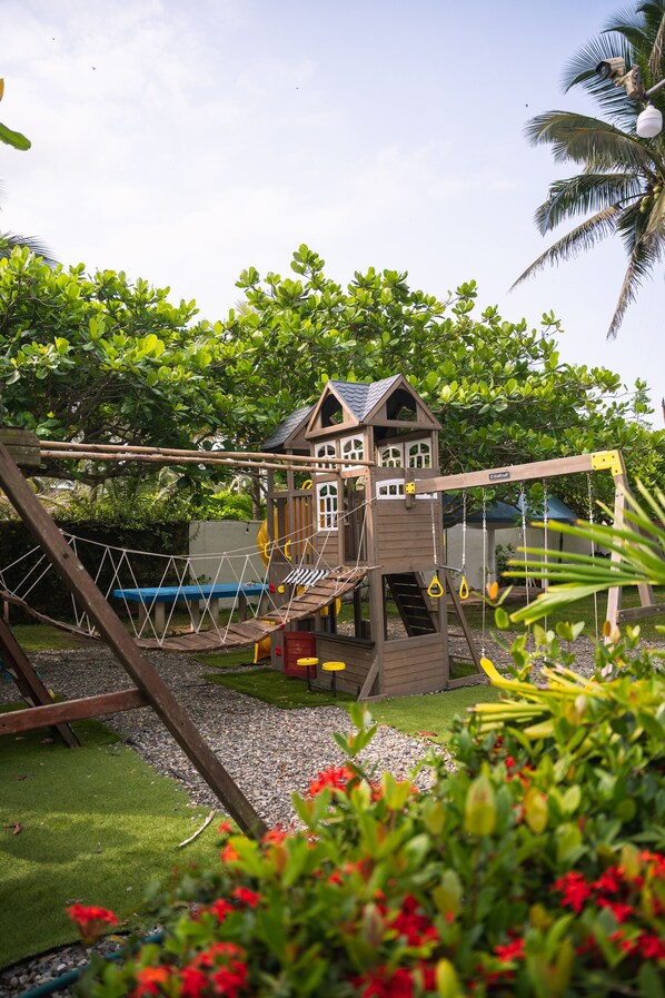 A wooden playhouse with a rope bridge and slide, surrounded by greenery and a clear sky. - Hotel Bungalows Marbella Costa Esmeralda (Tecolutla)
