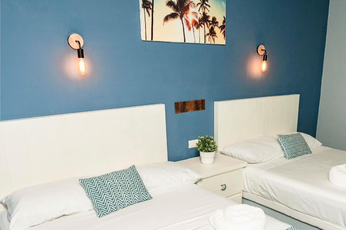 A hotel room with two beds, each with white linens and patterned pillows. There are wall-mounted light fixtures and a painting of palm trees.