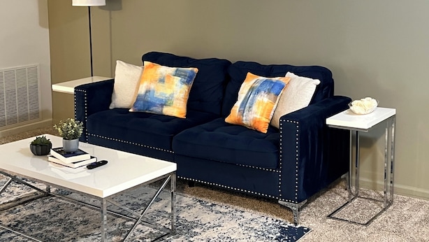 A dark blue sofa with decorative pillows, a white coffee table, a small side table with a white bowl, and abstract wall art.