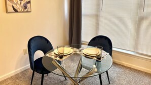 A dining area with a glass table, two black chairs, and a painting on the wall.