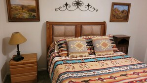 Een slaapkamer met een houten bed, beddengoed met een patroon, een lamp, een dressoir en twee ingelijste foto's aan de muur.