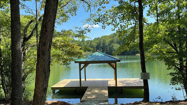 A wooden dock leading to a gazebo by a calm lake surrounded by trees.