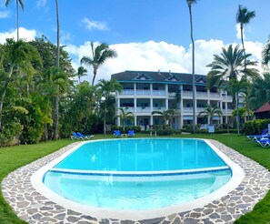 A swimming pool with a stone border, surrounded by palm trees and a building with balconies.