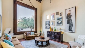 A cozy living room with a large window, a round coffee table, a sofa, a cabinet, and various framed pictures on the wall. - Midtown home with Pool Spa, fire pit, close to U of A, Restaurants, & Shopping (Tucson)