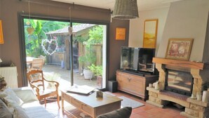 Una sala de estar con sofá, una mesa de centro, chimenea y televisor. Hay una puerta corrediza de vidrio que da a un patio con plantas y una terraza de madera.