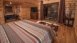 A log cabin bedroom with a large bed, a television on a wooden cabinet, a sliding glass door, and a cowhide rug.