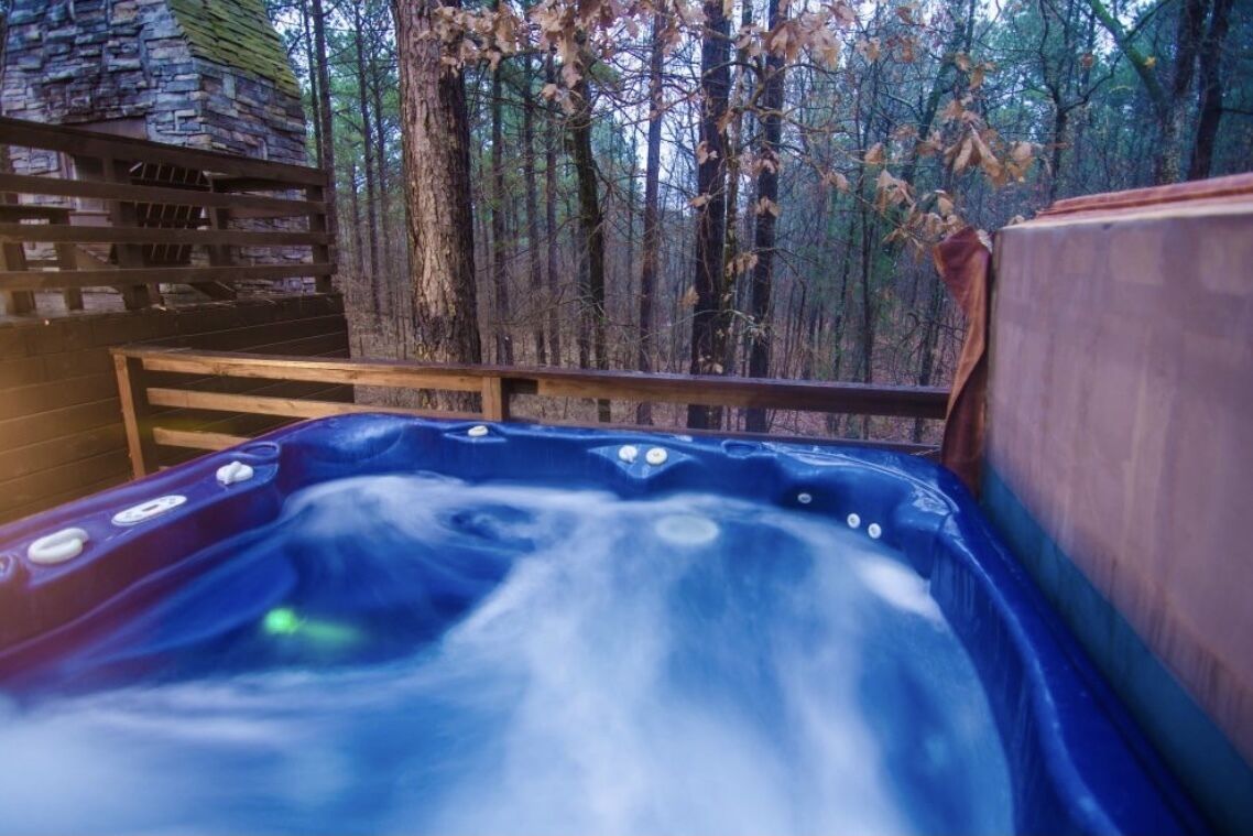 A hot tub surrounded by a wooden deck with a stone wall and a forest backdrop.