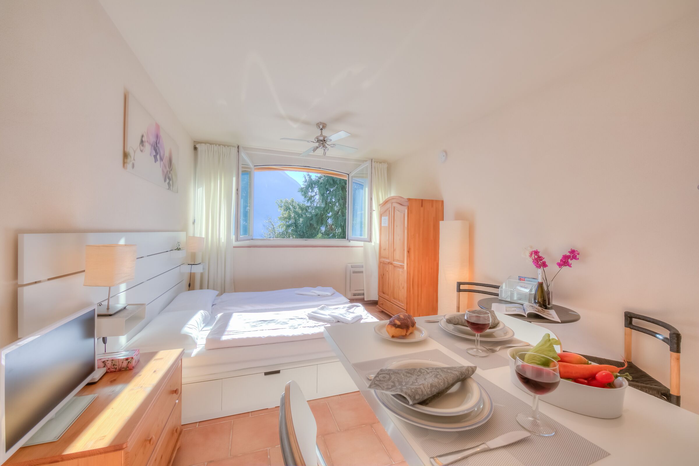 Quarto de hotel com uma cama, uma escrivaninha com um computador, uma mesa de jantar posta com pratos, copos e uma saladeira de frutas.