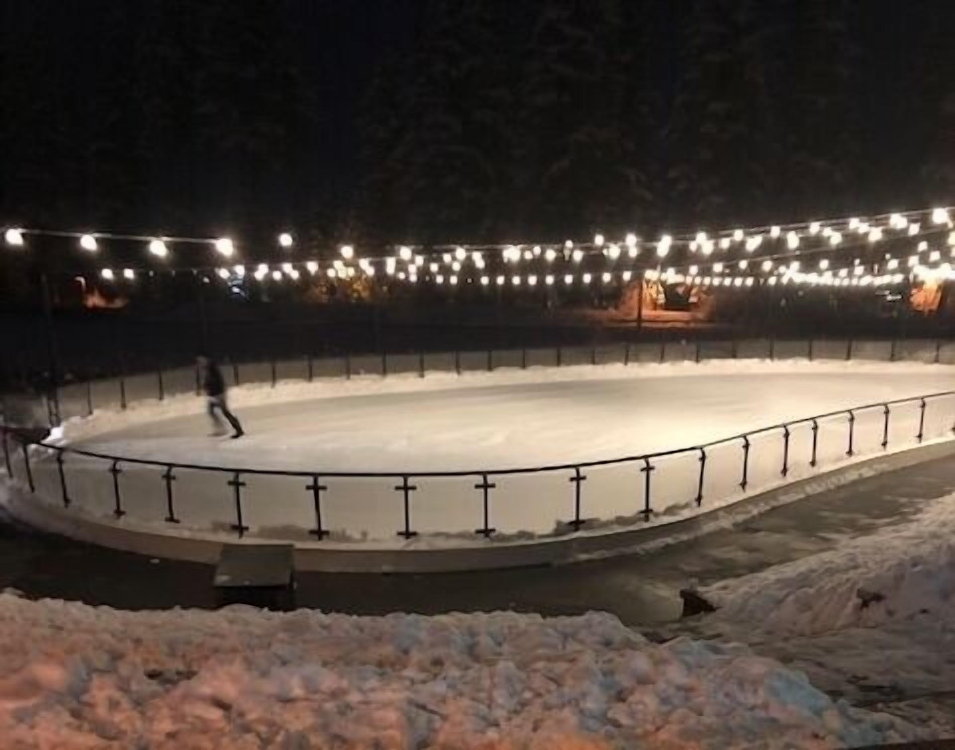 Eine Eislaufbahn im Freien bei Nacht mit Lichterketten darüber und einem verschneiten Vordergrund.