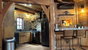 Una cocina rústica con vigas de madera, paredes de piedra, un refrigerador negro y una zona de bar con taburetes.