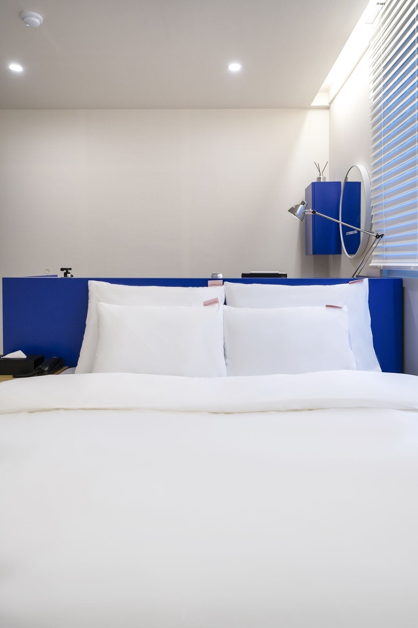 A modern hotel room with a neatly made bed, a blue headboard, a bedside table with a lamp, and a window with blinds.