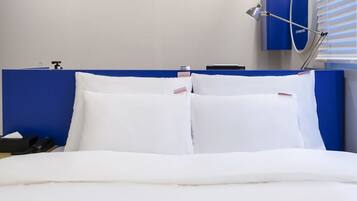 A modern hotel room with a neatly made bed, a blue headboard, a bedside table with a lamp, and a window with blinds.