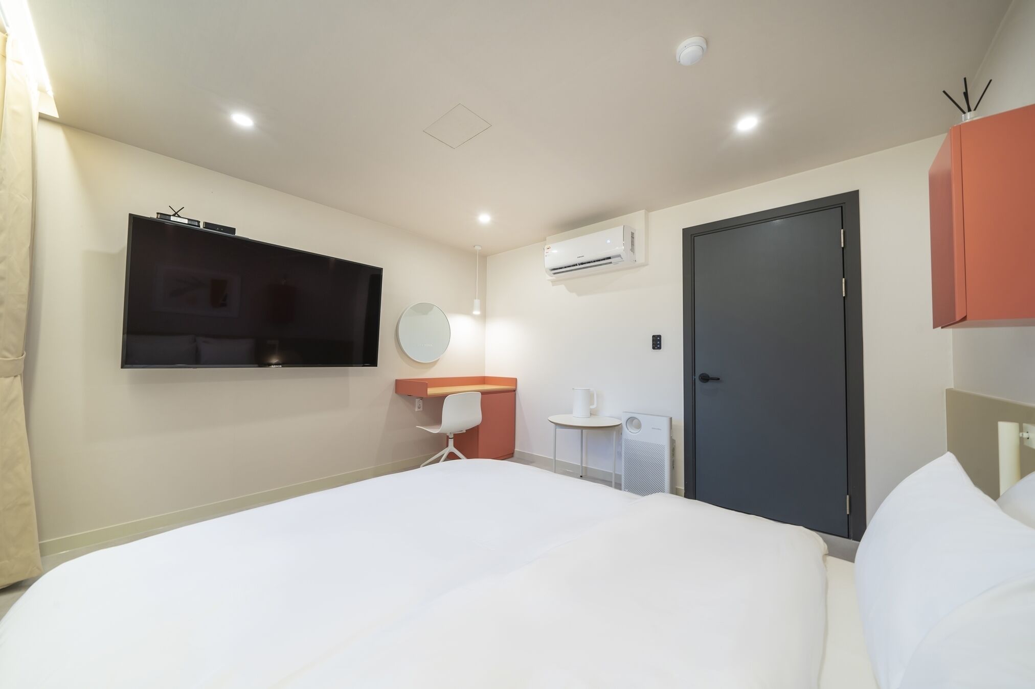 A hotel room with a bed, a desk with a chair, a mirror, an air conditioning unit, and a door.