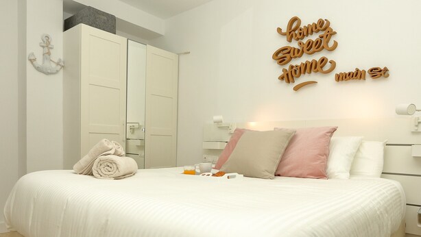 A bedroom with a large bed, a bedside table with a lamp, a wall-mounted mirror, and a decorative sign.