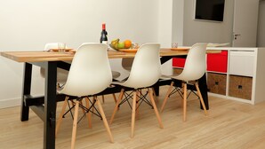 A dining area with a wooden table, white chairs, a fruit bowl, and a bottle of wine.