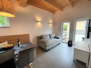 Una sala moderna con un sofá, una mesa de comedor y una pequeña cocina. Hay dos luces fijadas a la pared y una ventana que proporciona luz natural.