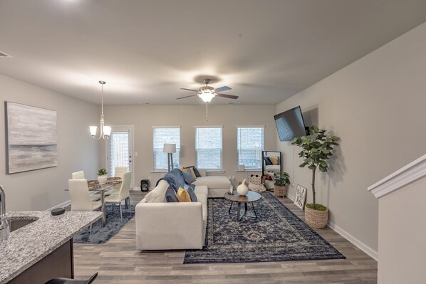 Un salon moderne doté d’un ventilateur de plafond, un coin repas avec une table et des chaises, et une cuisine avec un plan de travail en granit.