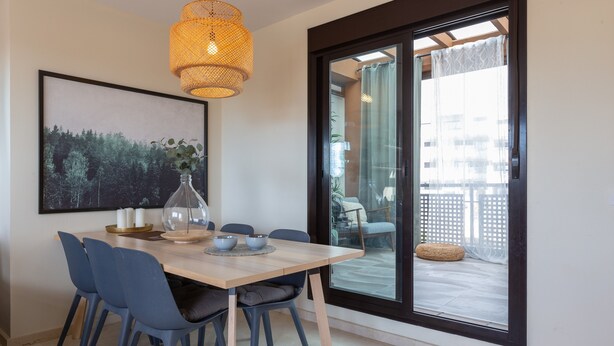 A dining area with a wooden table, blue chairs, a glass-top table, a woven pendant light, a framed landscape painting, and a sliding glass door leading to an outdoor balcony. - Vacation Home Penthouse La Cala De Mijas with Sea View, Pool (La Cala de Mijas)