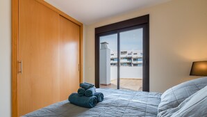 A bedroom with a bed, a wooden wardrobe, a stack of rolled towels, and a view of buildings through a glass door. - Vacation Home Penthouse La Cala De Mijas with Sea View, Pool (La Cala de Mijas)