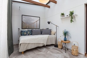 Une chambre à coucher avec un lit, une table de chevet avec une lampe, un tableau encadré au mur et une petite étagère avec des plantes.