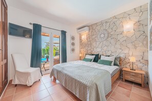 A bedroom with a bed, a chair, a nightstand, a TV, and a stone wall.