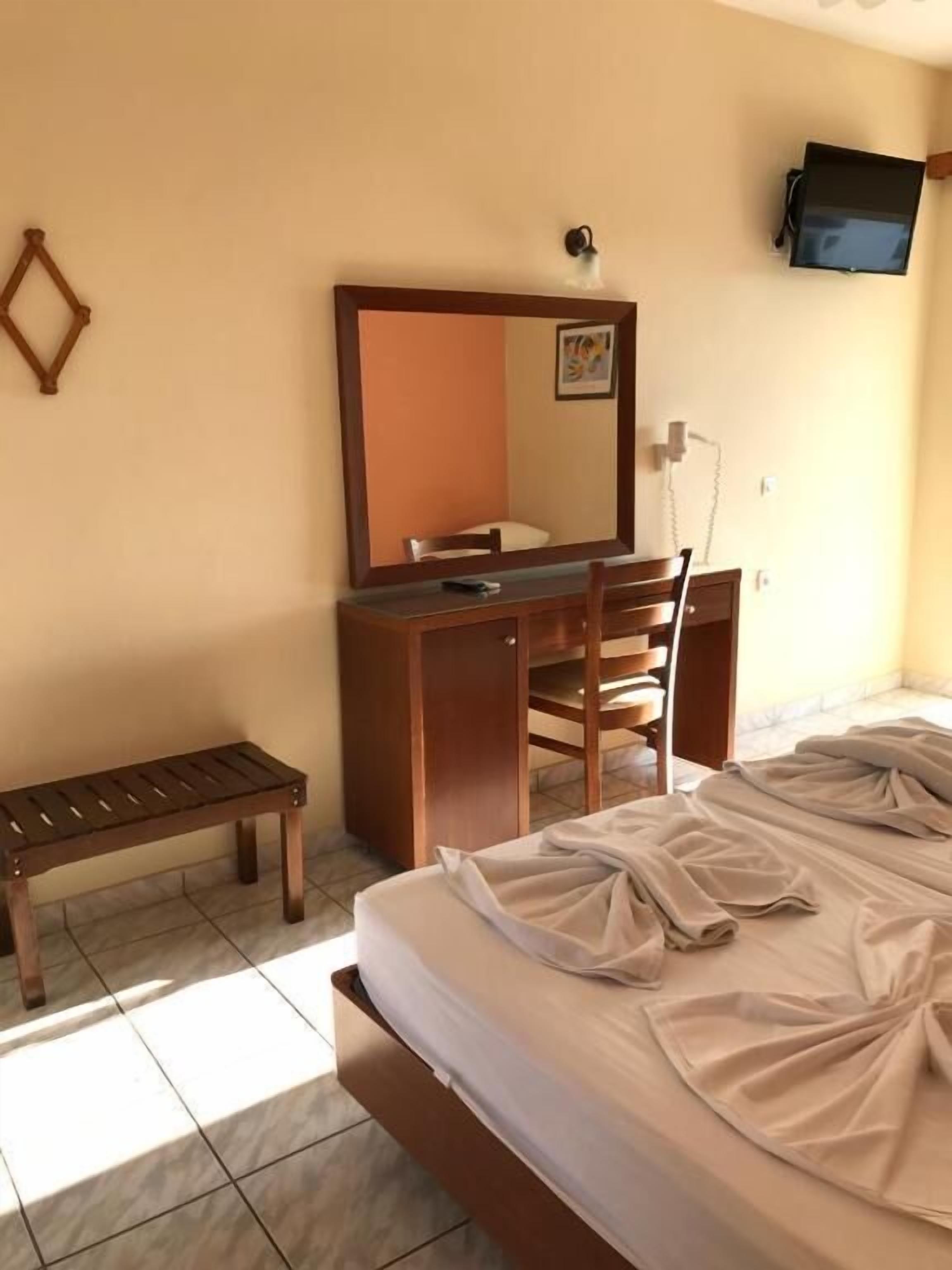 A bedroom with a bed, a wooden desk, a chair, a bench, a wall-mounted TV, and a mirror.
