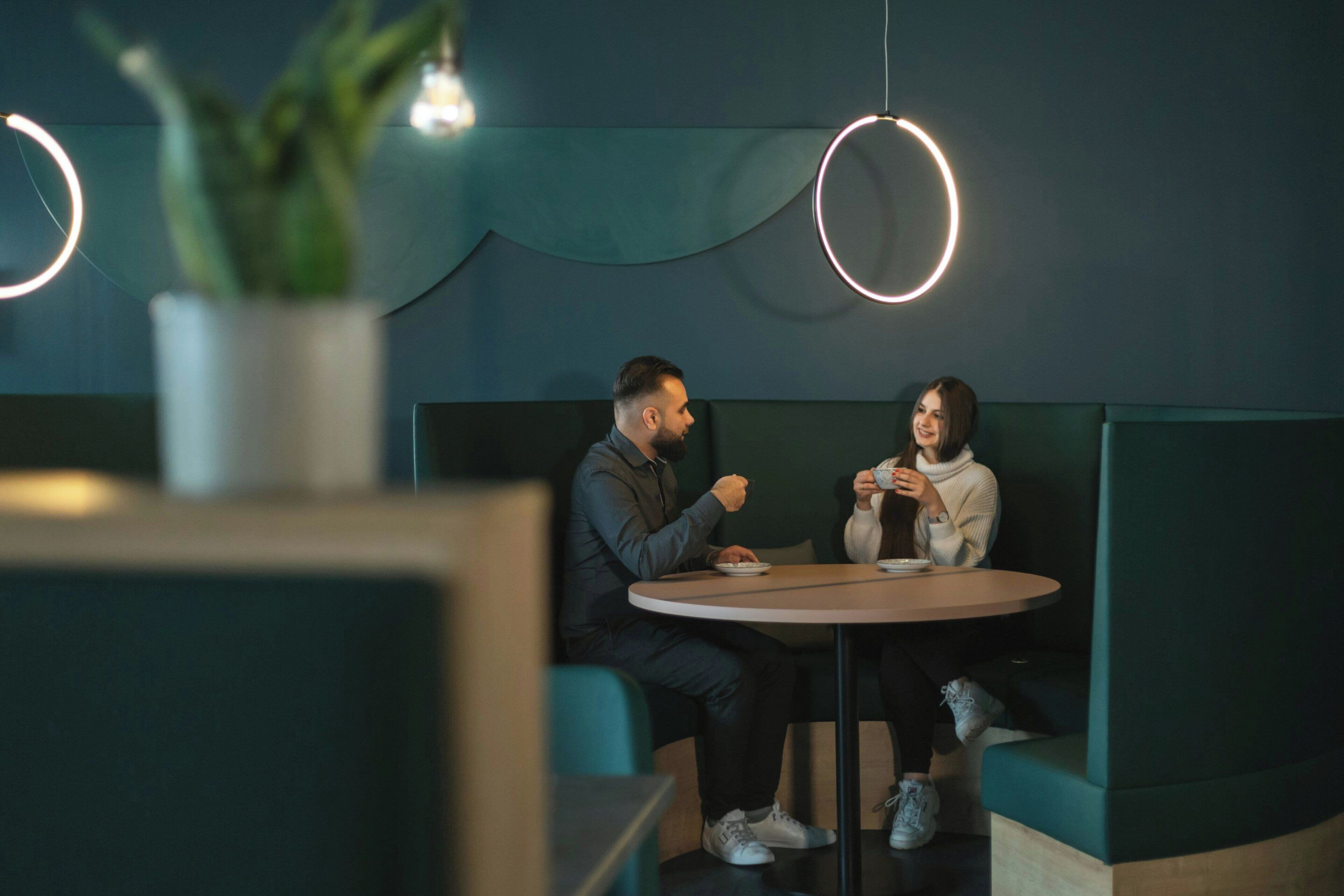 Deux personnes sont assises autour d’une table ronde dans un intérieur moderne aux accents turquoise et doté de luminaires circulaires.
