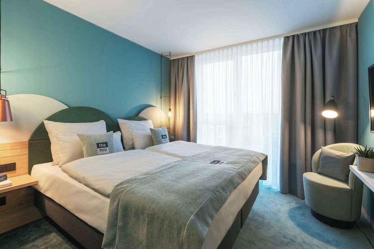 Habitación de hotel con cama, mesitas de noche, una silla, televisor y ventana con cortinas.