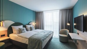 Una habitación de hotel moderna con cama, mesita de noche, lámpara, silla, televisor y ventana grande con cortinas.