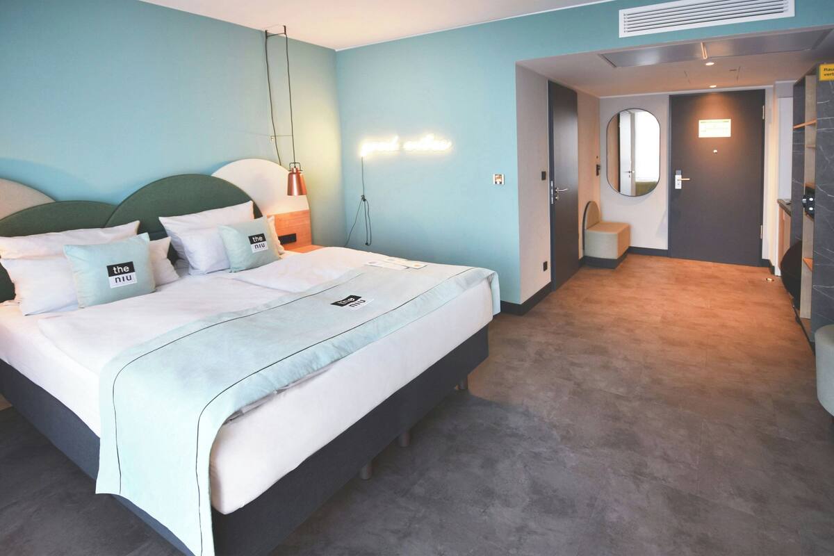 A hotel room with a bed, bedside table, lamp, wall-mounted light, mirror, and a door leading to a bathroom.